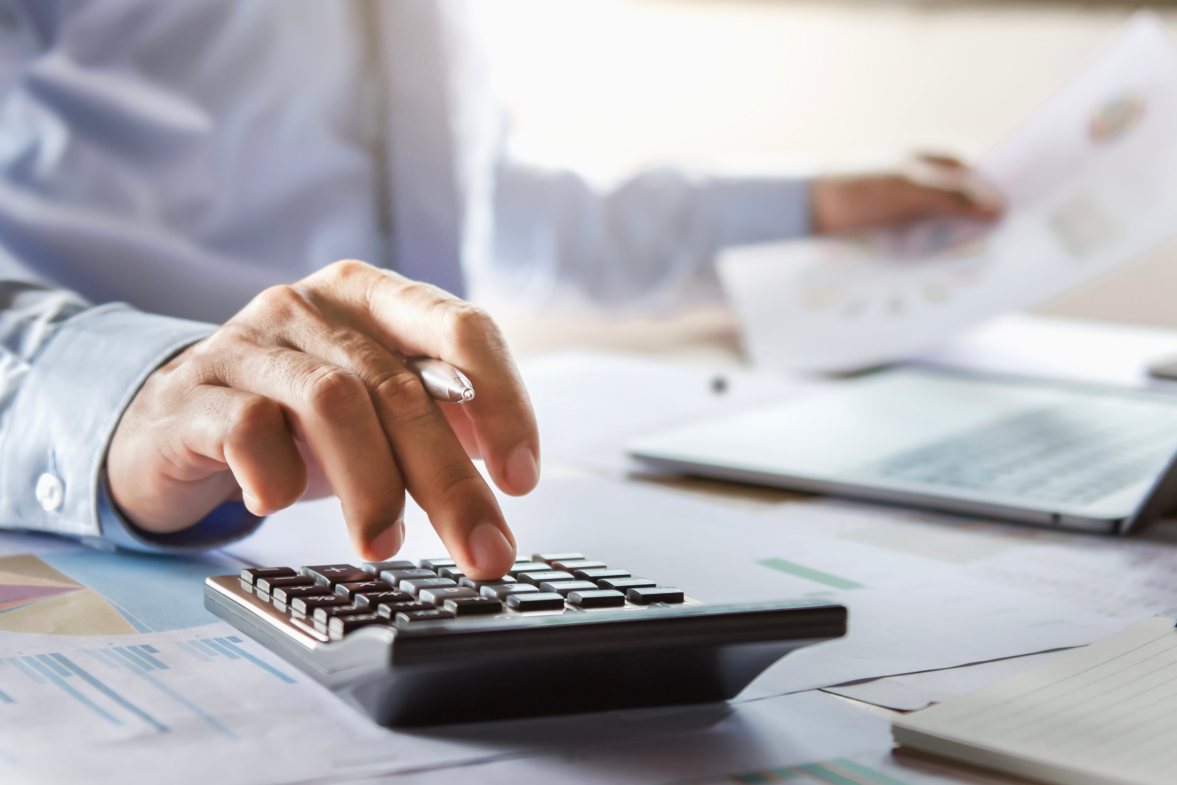 Person in a blue shirt using a calculator at a desk with papers and a laptop.