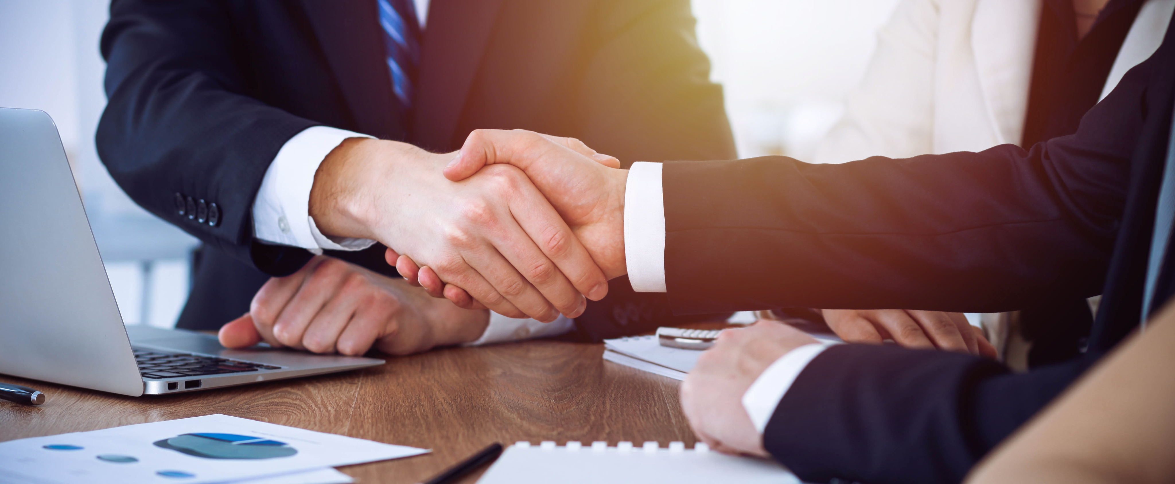 Two people in business attire shaking hands over a table with documents and a laptop.