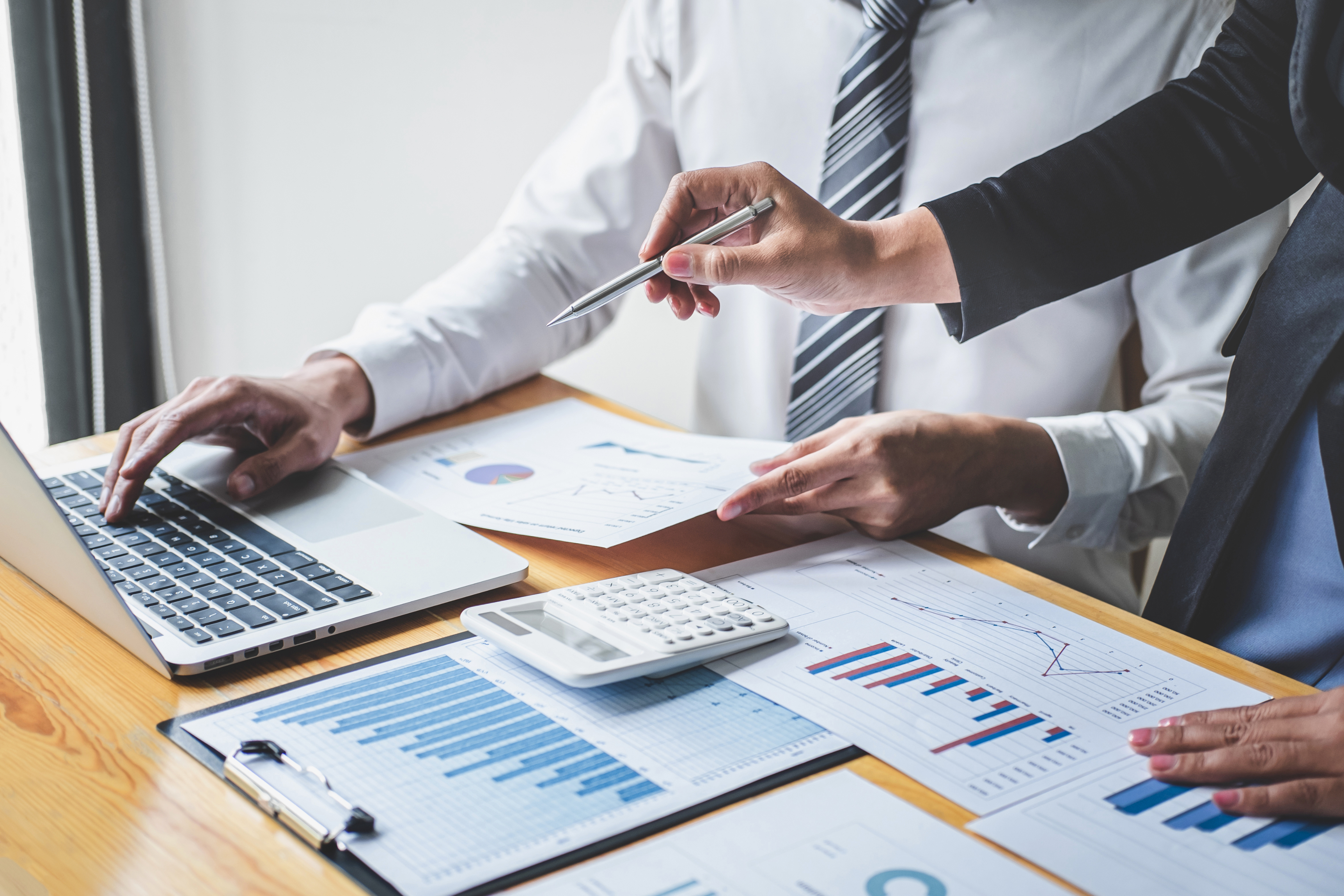 Two people work at a desk with a laptop, calculator, and various financial charts and graphs on paper.