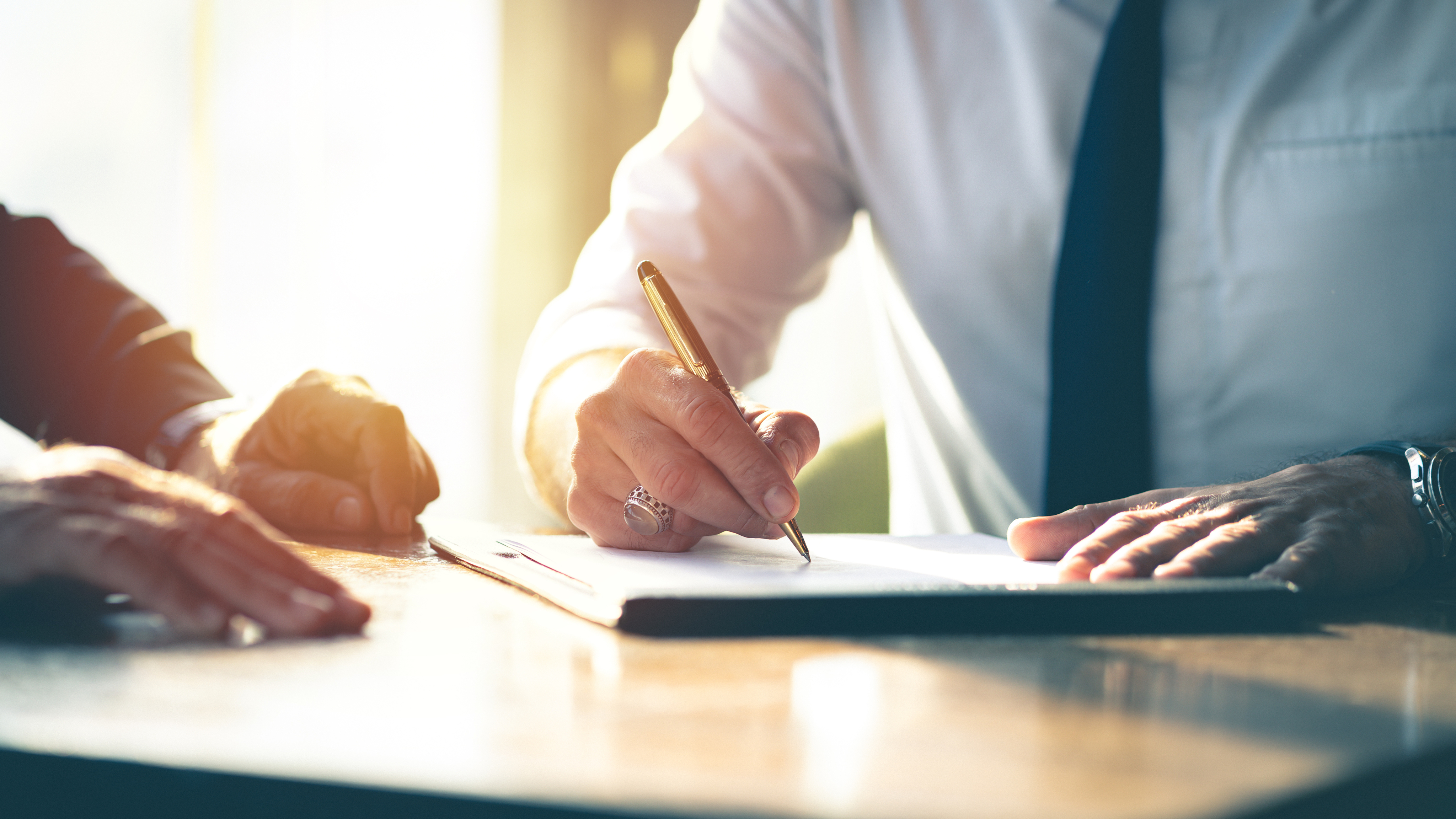 A person in a suit signs a document on a desk with a pen, while another hand rests nearby.