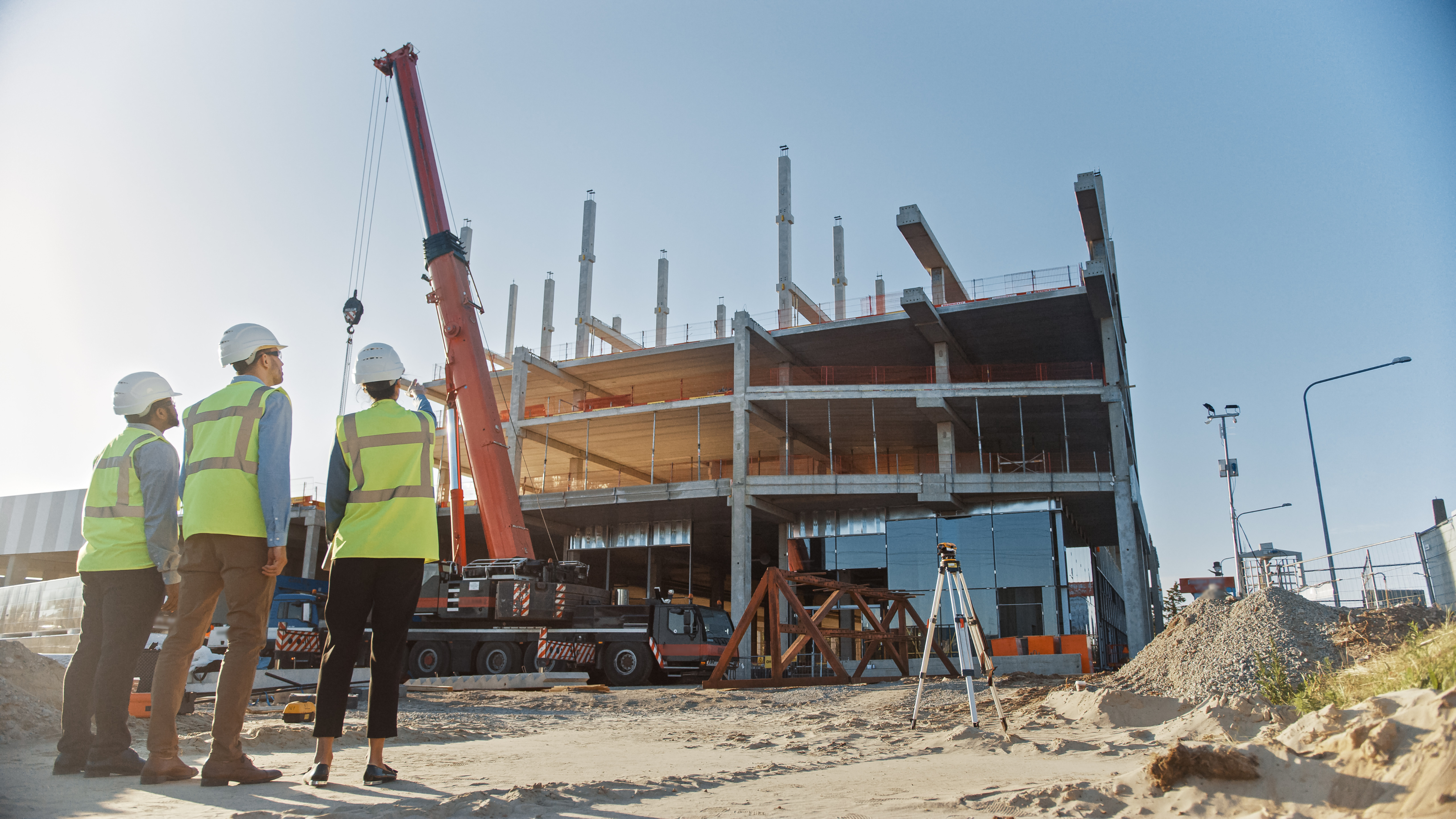 Three construction workers wearing safety gear observe a building under construction with a crane in the background on a clear day.