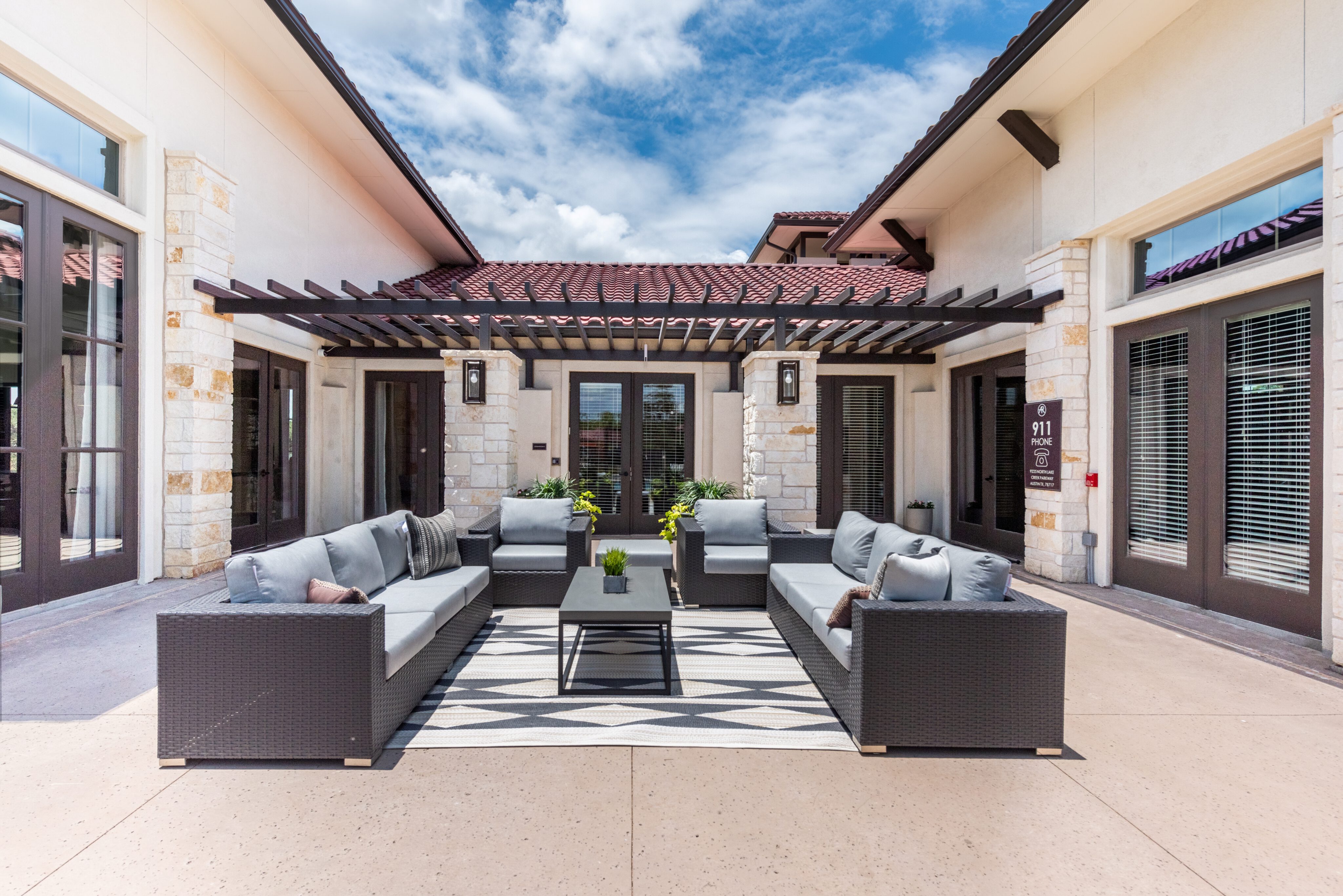 Outdoor patio area with wicker sofas and a coffee table under a pergola. Neutral tones complement the building's architecture. Sky is partly cloudy.