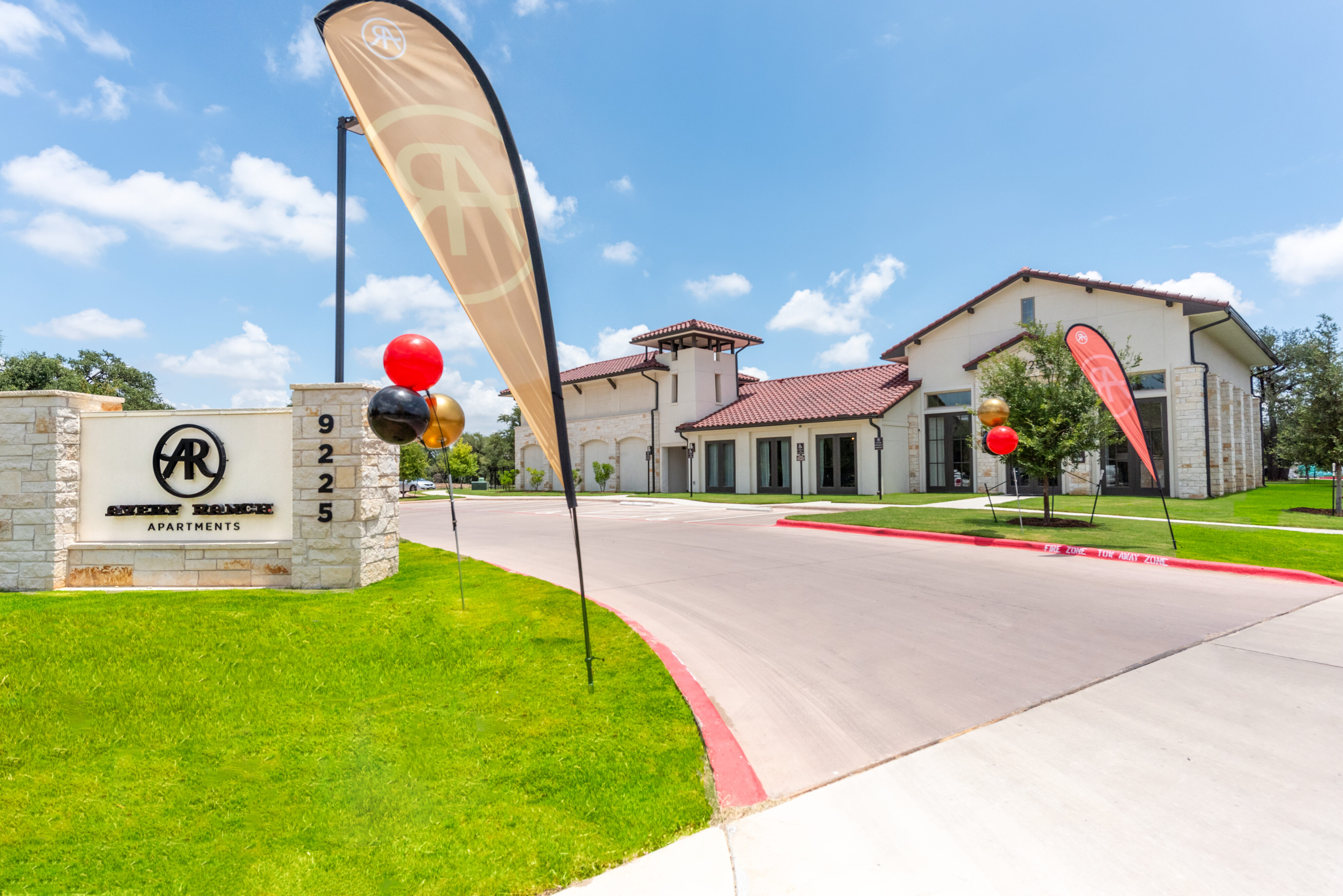 The image shows a modern apartment complex with a sign, flags, and balloons at the entrance. The sky is clear and blue.