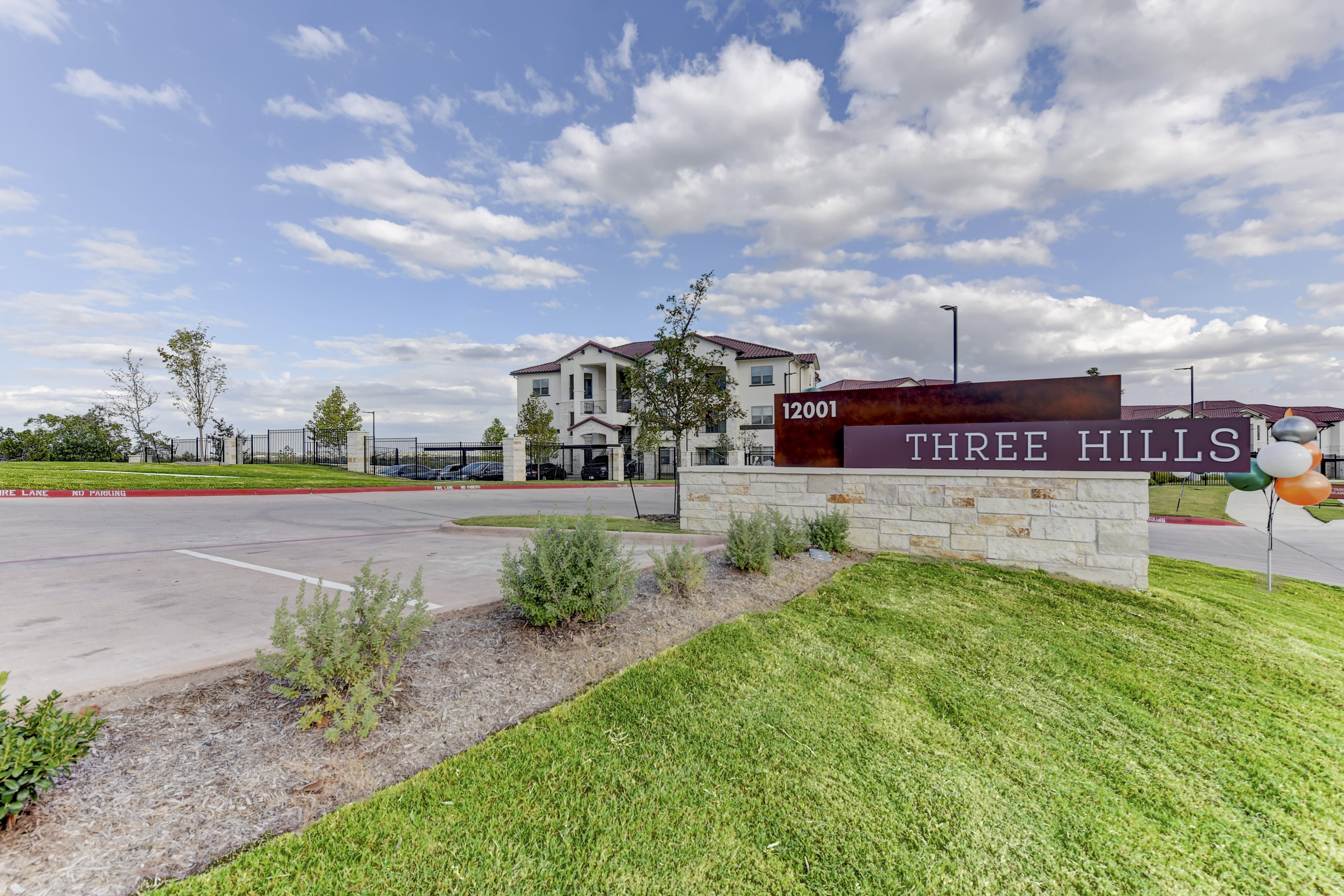 Entrance to Three Hills residential complex with a large sign, pavement, and neatly trimmed grass under a partly cloudy sky.