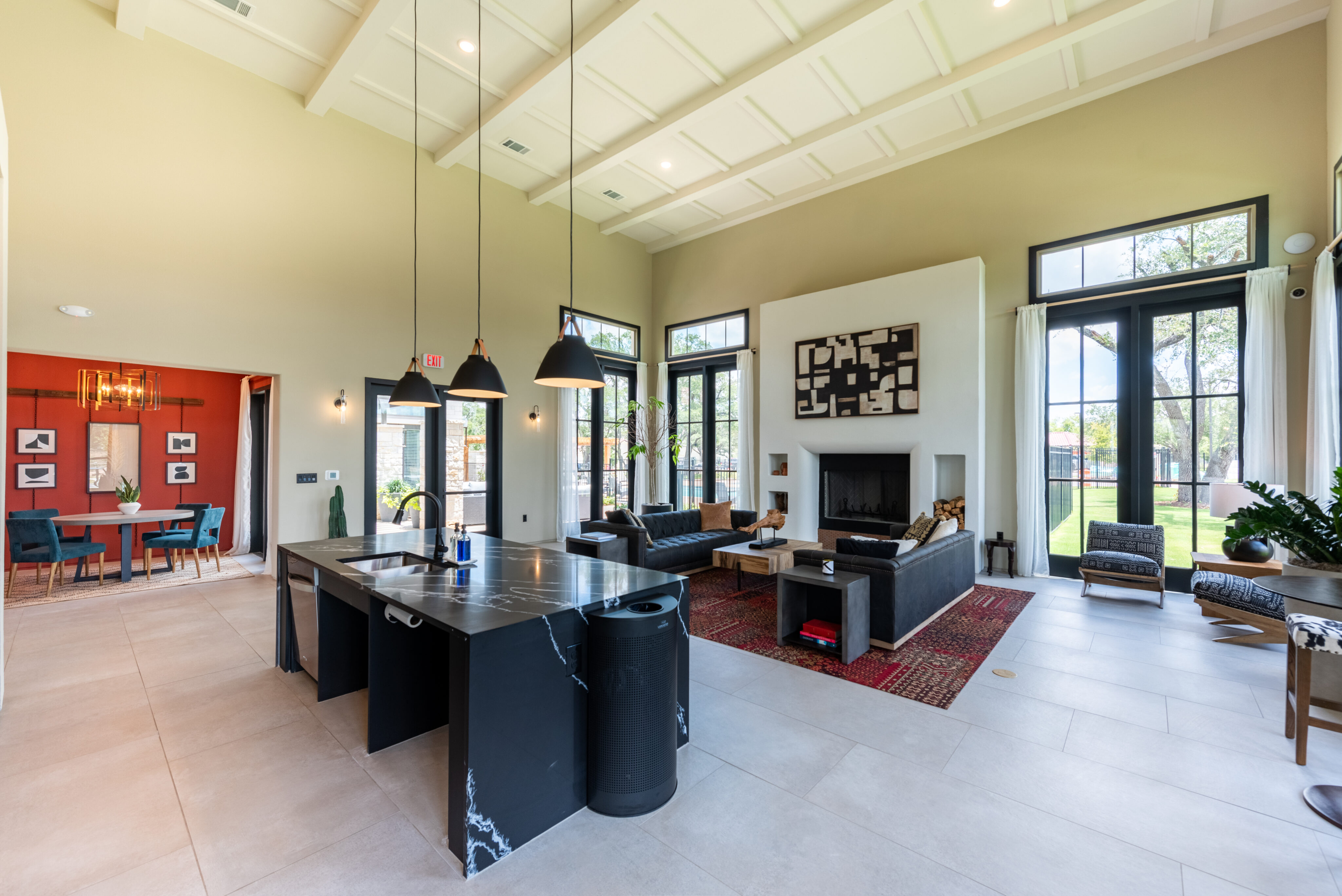 Spacious modern living area with a kitchen island, black countertops, and pendant lights. Seating area with couches, a fireplace, large windows, and an adjoining dining room in the background.