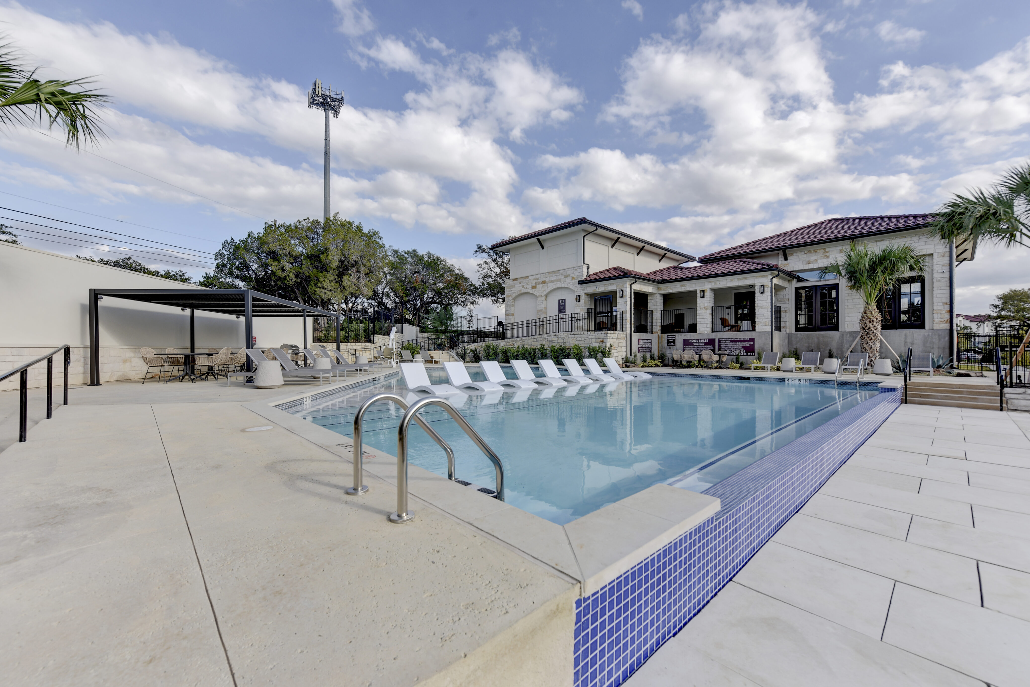 Outdoor swimming pool with lounge chairs and palm trees. A two-story building with a covered patio is in the background. The sky is partly cloudy.