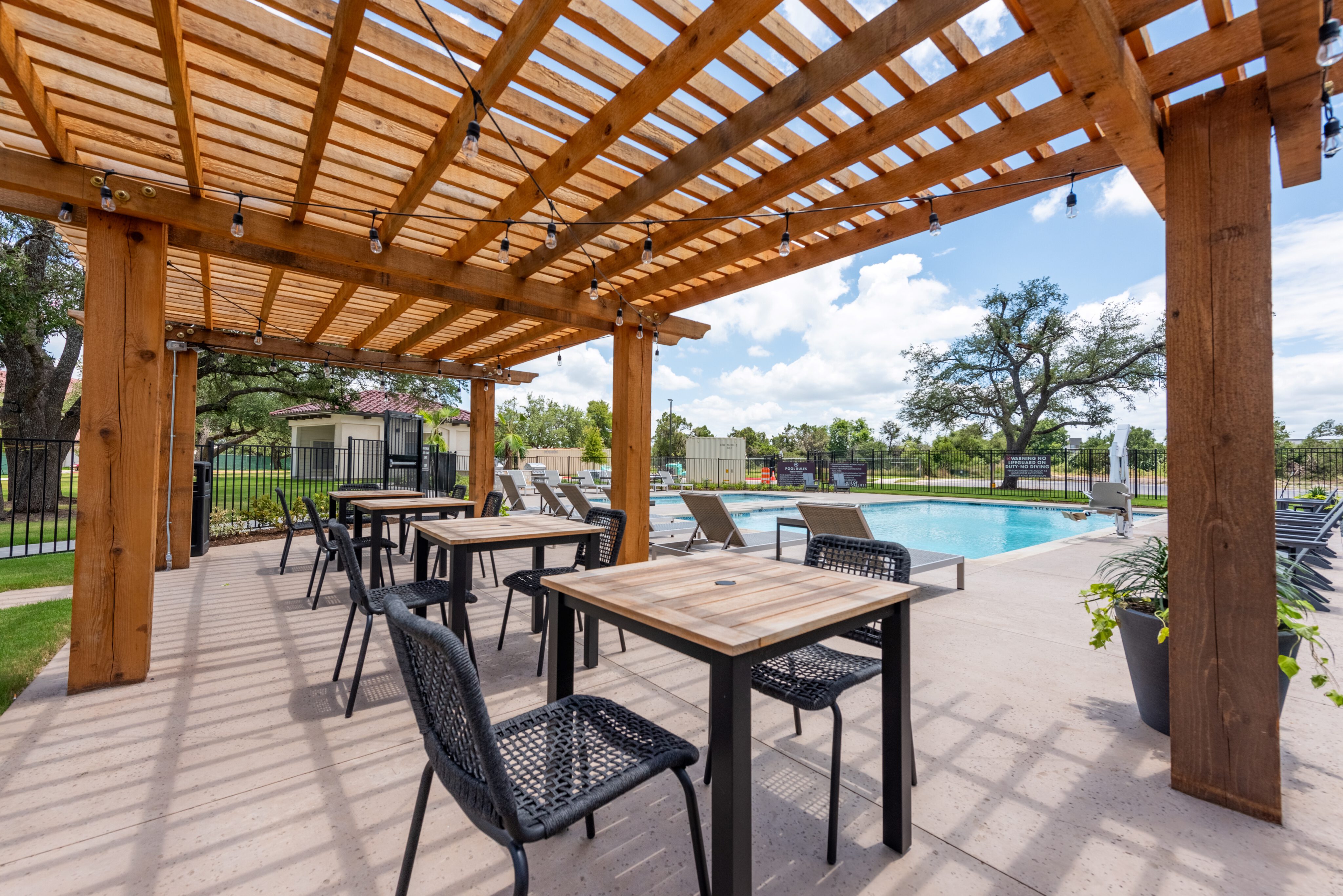 Outdoor patio with wooden pergola and string lights, featuring tables, chairs, and a view of a swimming pool with lounge chairs.