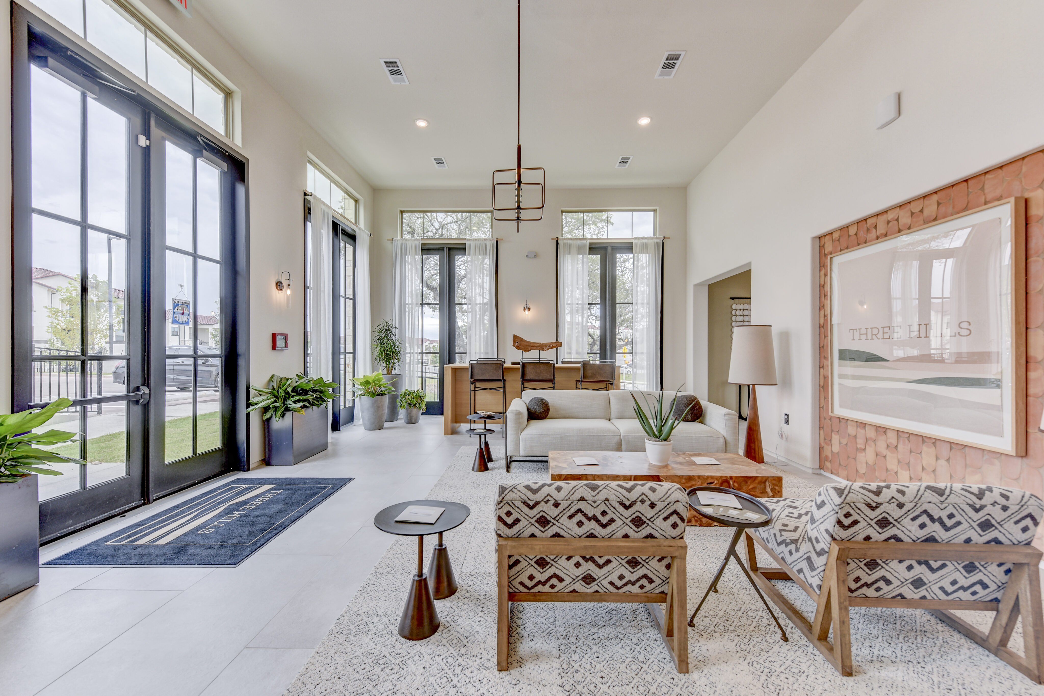 Bright, modern living room with patterned furniture, large windows, and a view of greenery outside. Decor includes a rug, plants, artwork, and a multi-bulb pendant light.