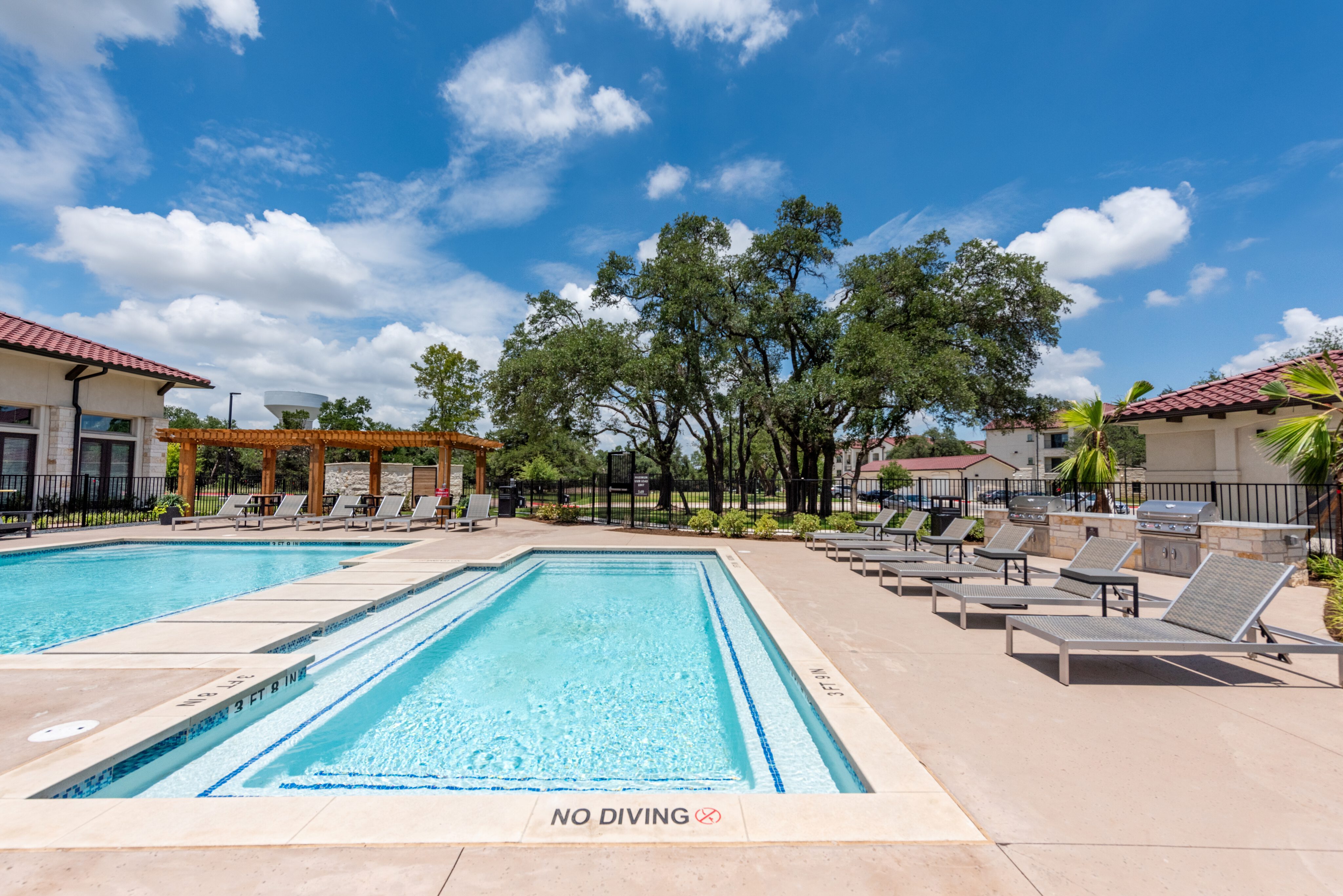 Outdoor swimming pool with lounge chairs, a pergola, and trees in the background under a blue sky with clouds.