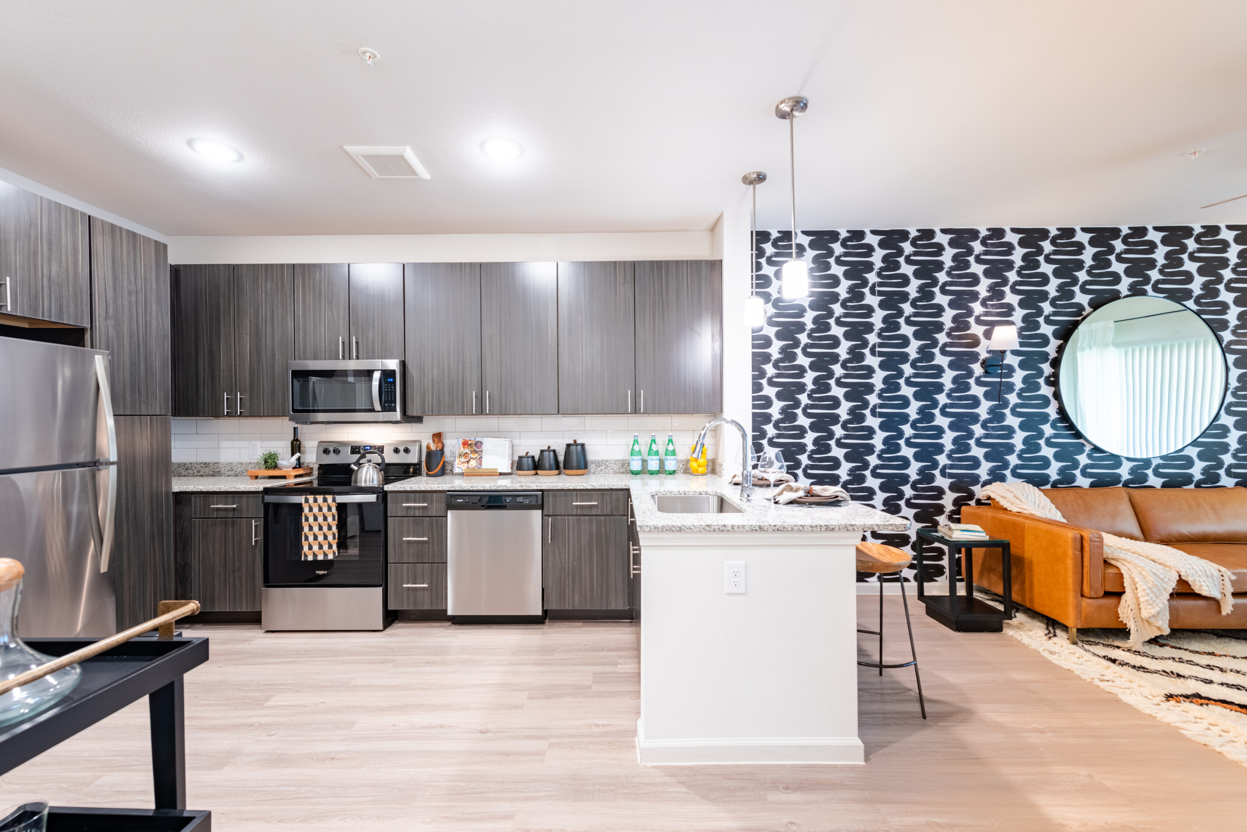 Modern kitchen and living area with stainless steel appliances, gray cabinets, patterned wallpaper, a round mirror, and a tan leather sofa.