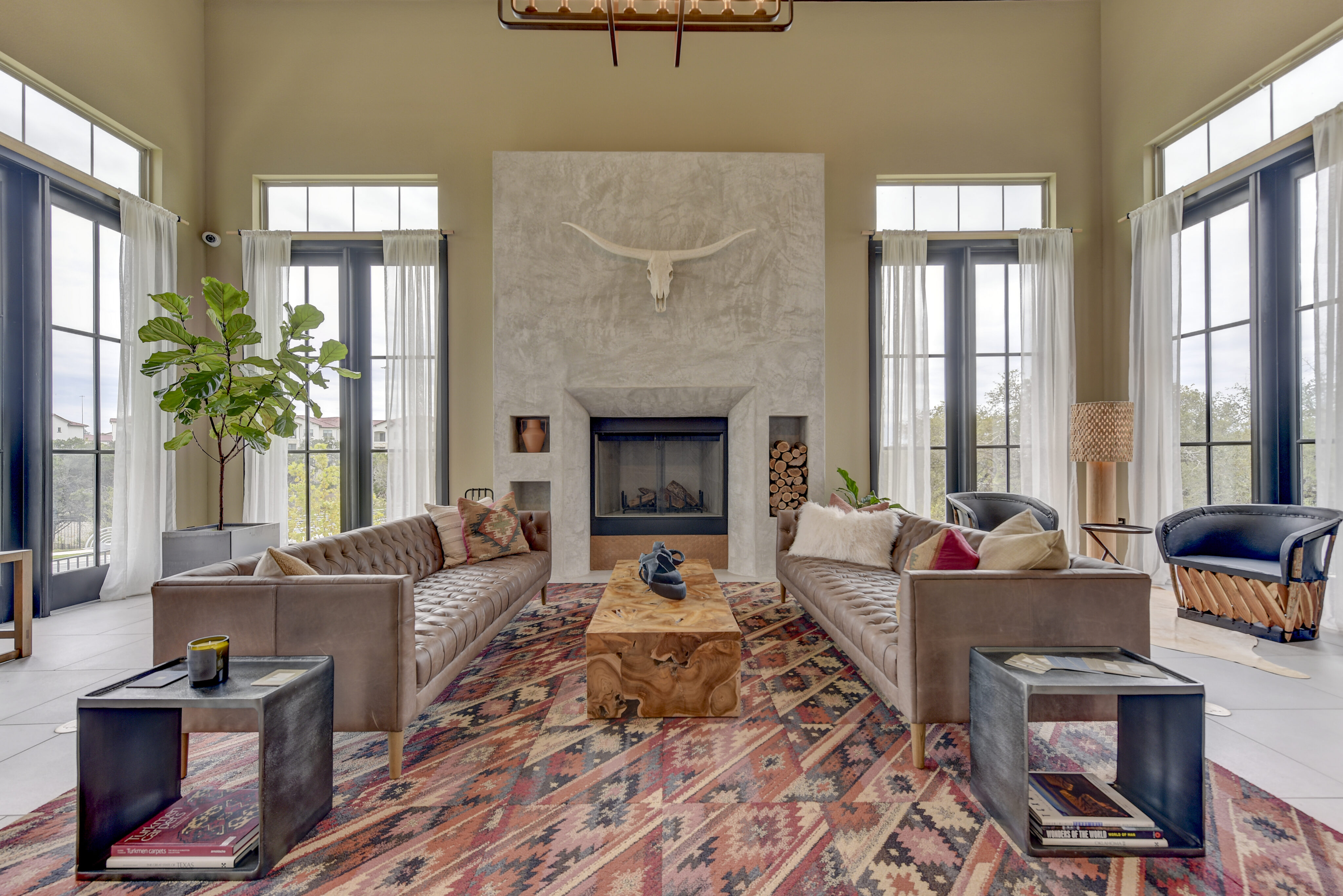 Modern living room with two leather sofas facing each other, a central wooden coffee table, a fireplace with a decorative skull, a large plant, and floor-to-ceiling windows.