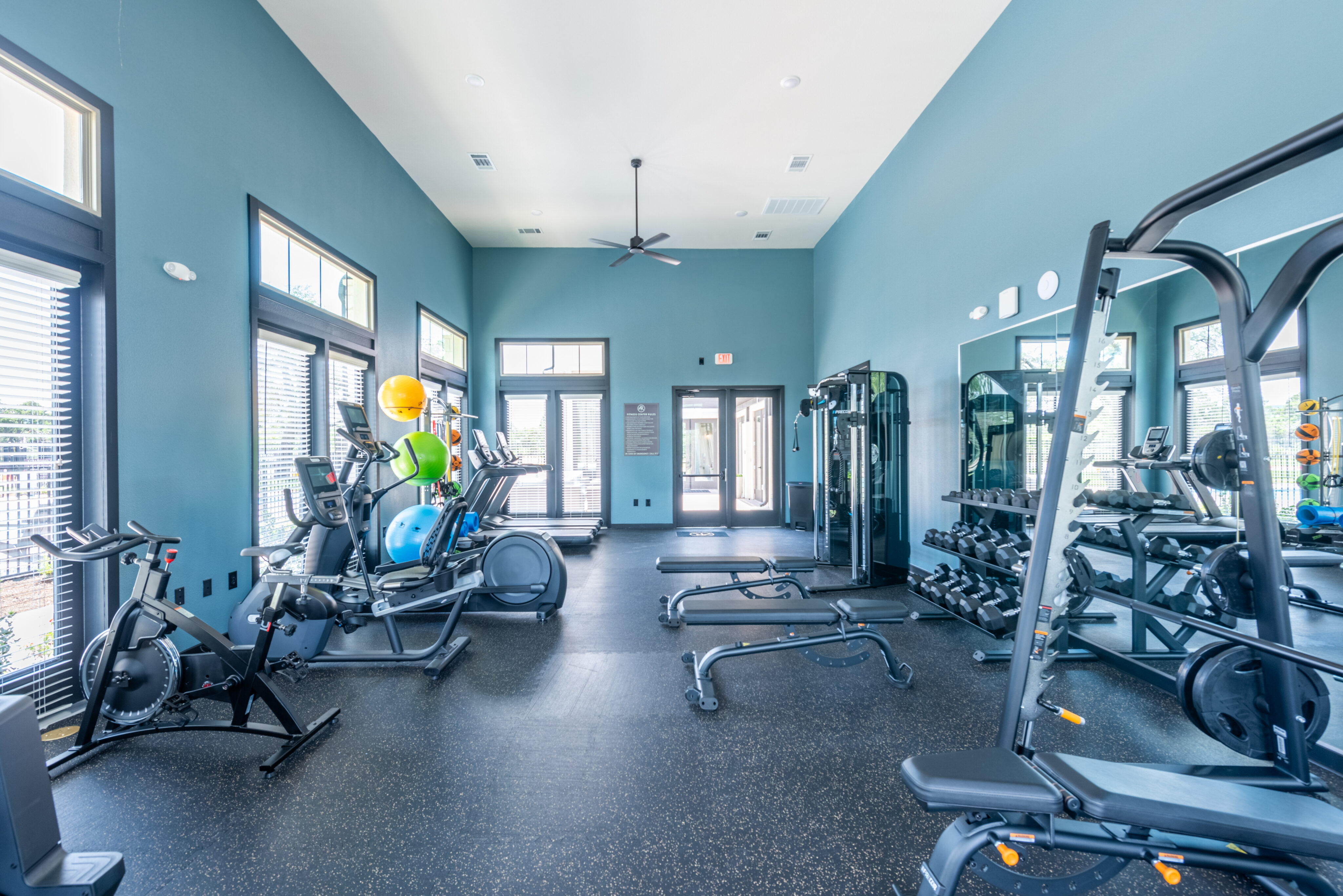 A bright, spacious gym with equipment like treadmills, weights, and balance balls, featuring large windows and a black rubber floor.