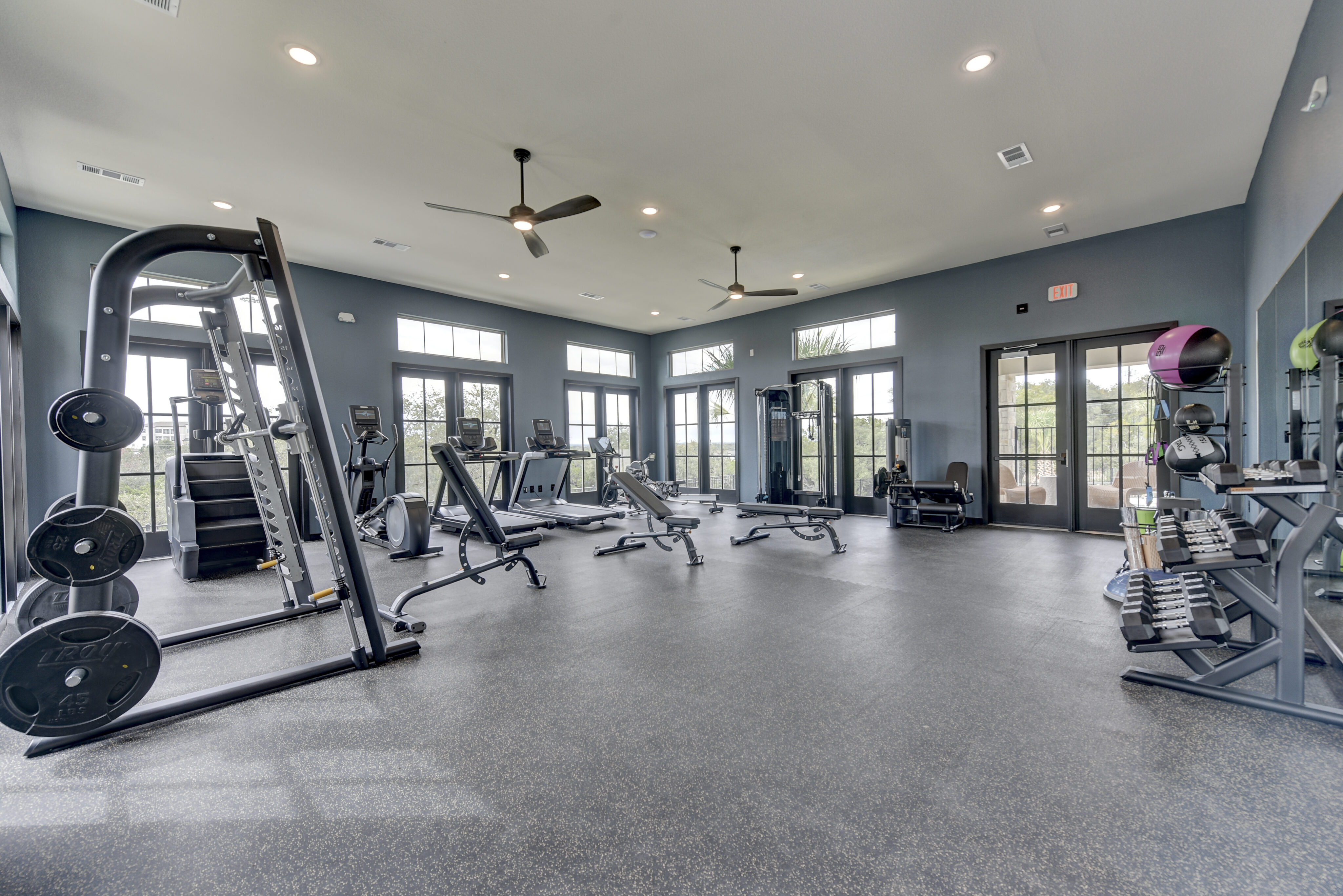 Spacious gym with various exercise equipment, including treadmills, weight machines, and dumbbells. Large windows and ceiling fans provide natural light and ventilation.