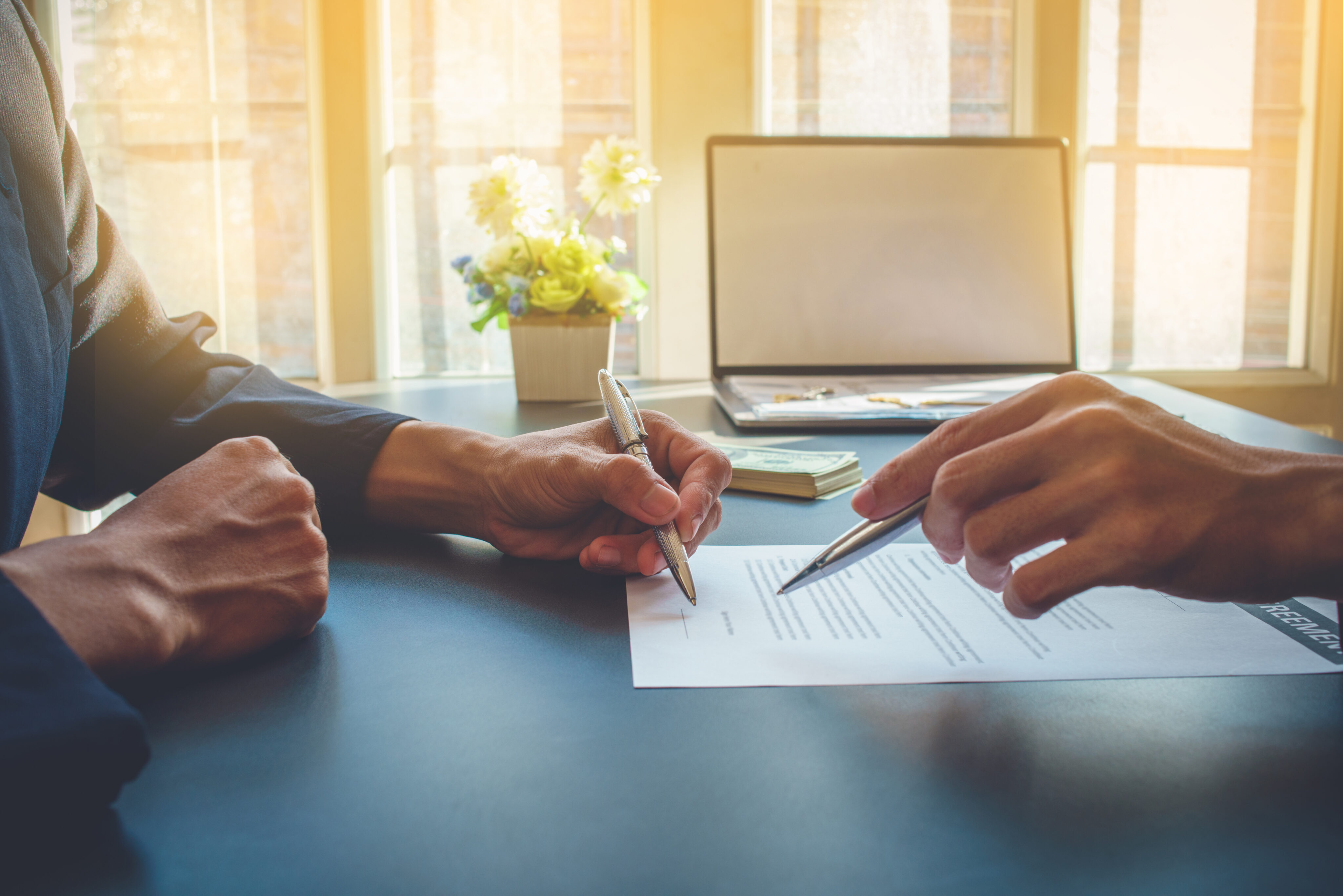 Two people sitting at a table, one signing a document with a pen. An open laptop, flowers, and a stack of books or papers are in the background.