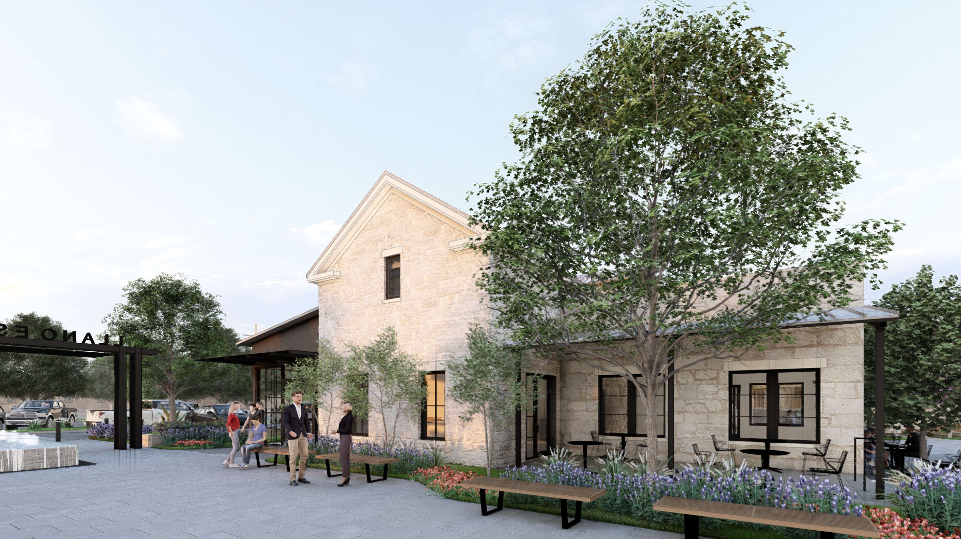People walk and gather outside a stone building with large windows, surrounded by trees, benches, and flower beds on a paved area.